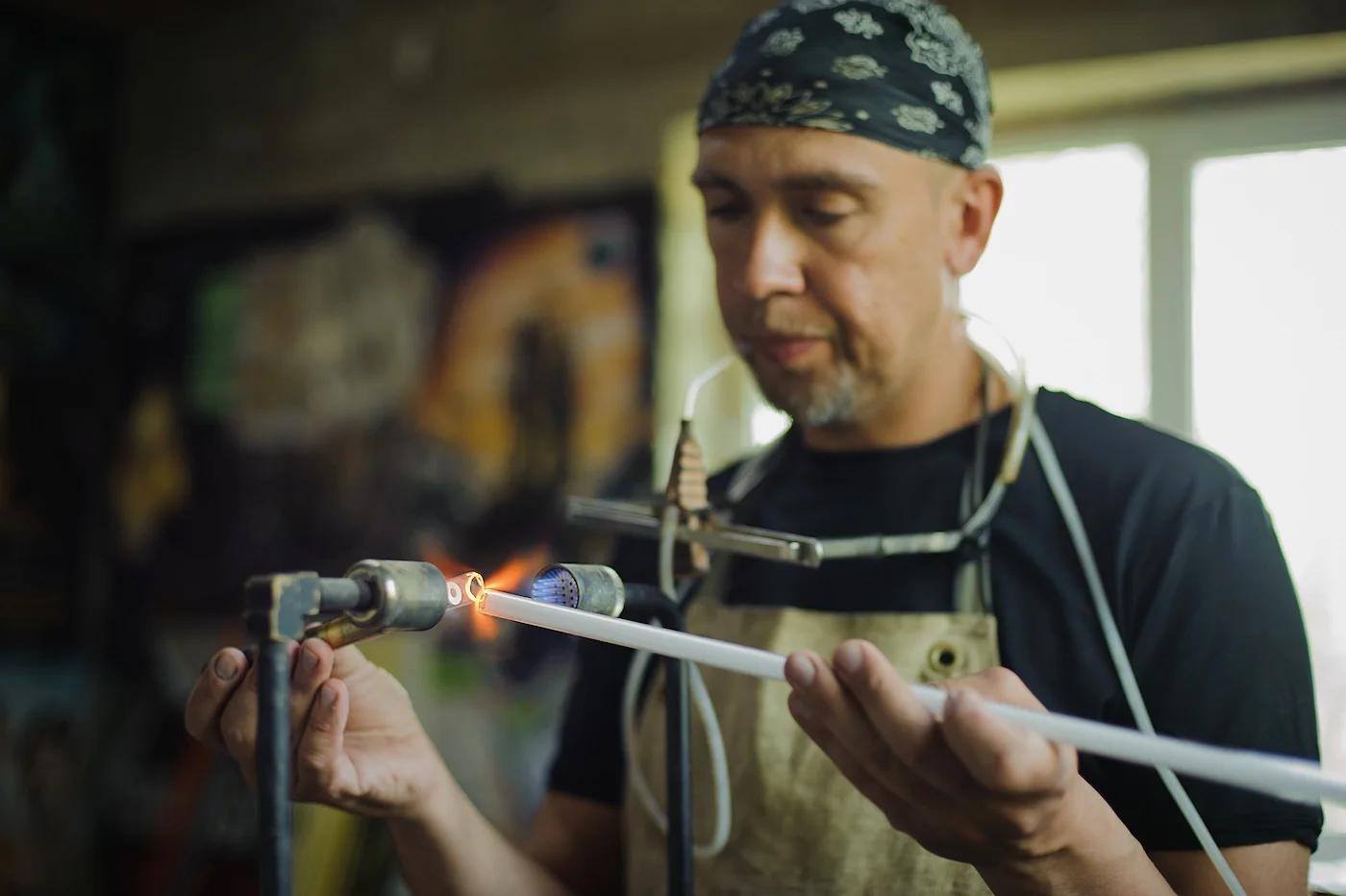 Craftsman in a workshop using a blowtorch to shape a glass tube for a neon sign. He is focused on the task, wearing a bandana and an apron, showcasing the intricate process of creating custom neon signage.