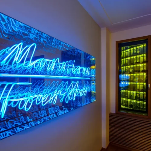 Modern neon sign in blue cursive, displayed in a glass frame on a wall, creating a mirror effect with multiple reflections. The room has a warm lighting ambiance.