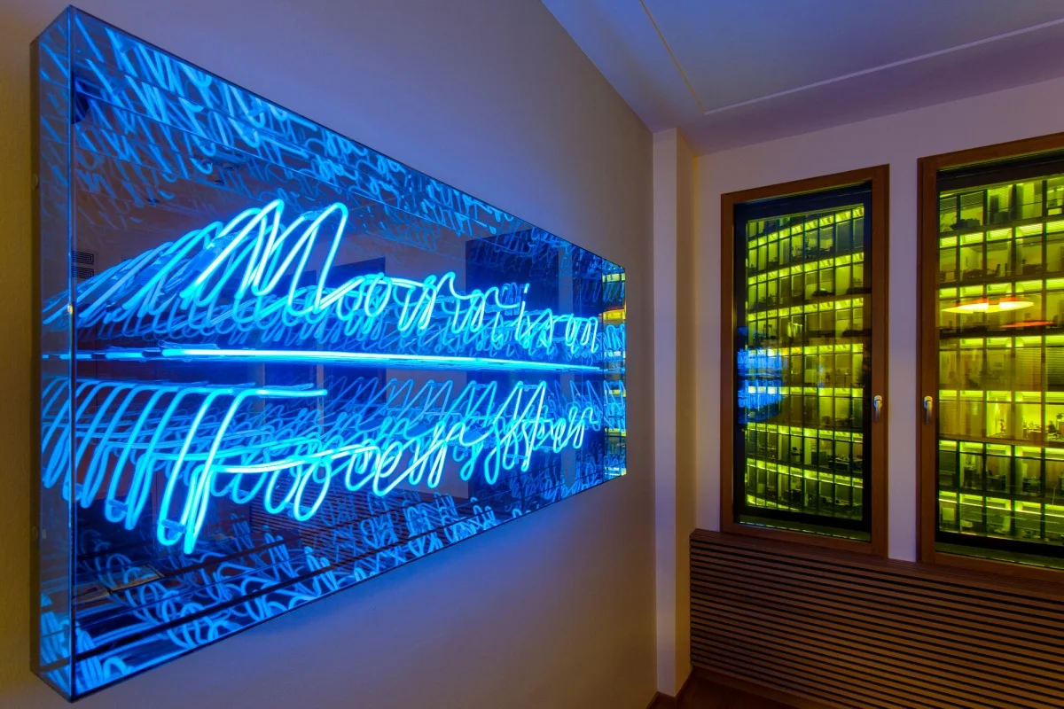 Modern neon sign in blue cursive, displayed in a glass frame on a wall, creating a mirror effect with multiple reflections. The room has a warm lighting ambiance.