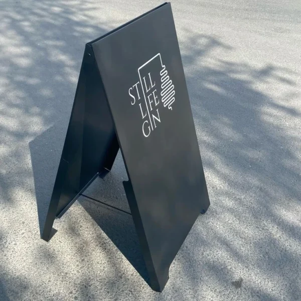 Black A-frame sidewalk sign for "Still Life Gin" displayed outdoors. The sign features white text and a minimalist line drawing of a gin still, creating a modern and sophisticated look. Ideal for promoting business and event specials.