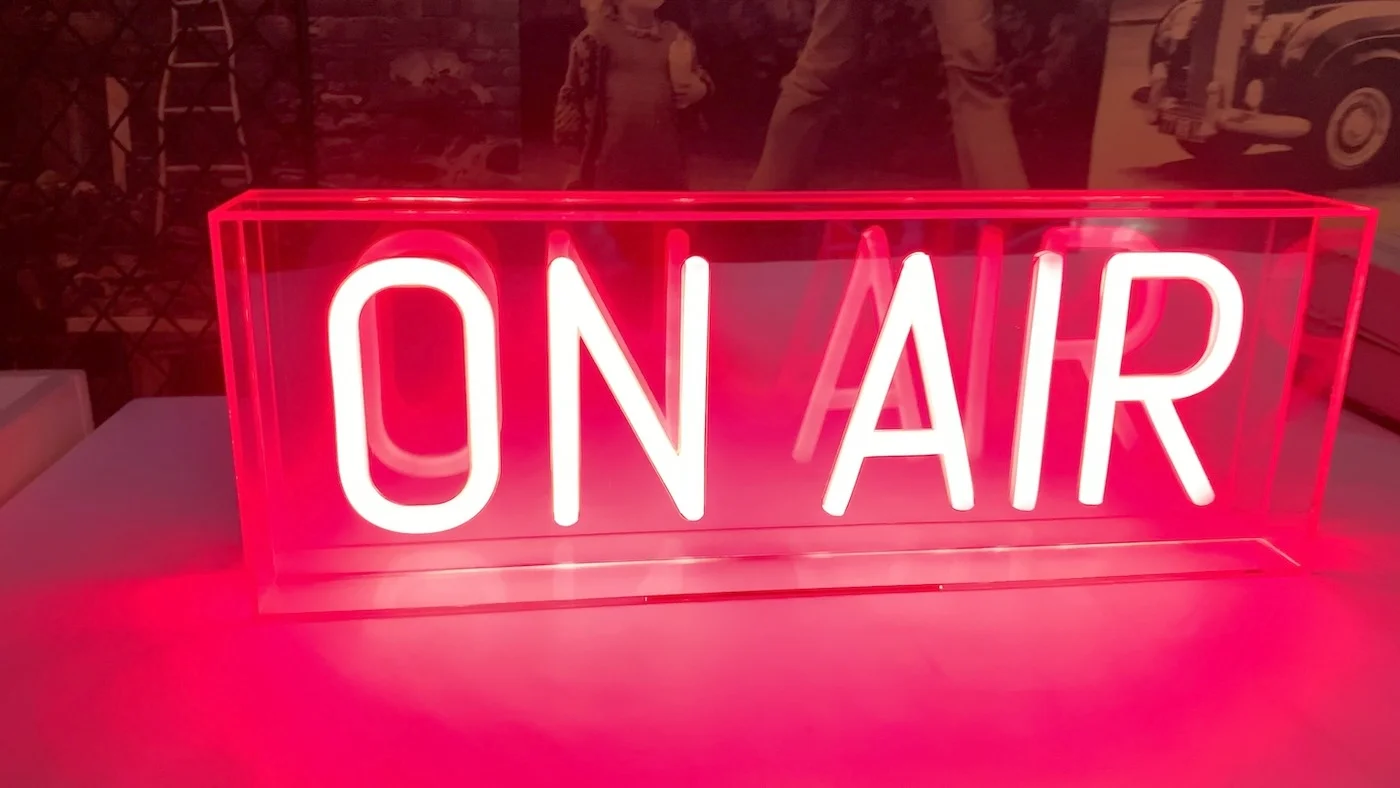 Neon sign with the phrase "ON AIR" in white letters, encased in a clear acrylic box with a bright red glow. The sign is positioned on a table, creating a vibrant and eye-catching display suitable for a recording studio or broadcasting environment.