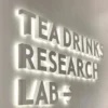 A modern wall sign with illuminated white 3D letters reads "TEA DRINKS RESEARCH LAB" against a light-colored background. The letters are bold and evenly spaced, casting subtle shadows and creating a striking visual effect.