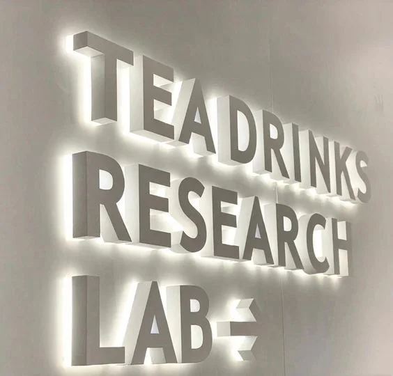 A modern wall sign with illuminated white 3D letters reads "TEA DRINKS RESEARCH LAB" against a light-colored background. The letters are bold and evenly spaced, casting subtle shadows and creating a striking visual effect.