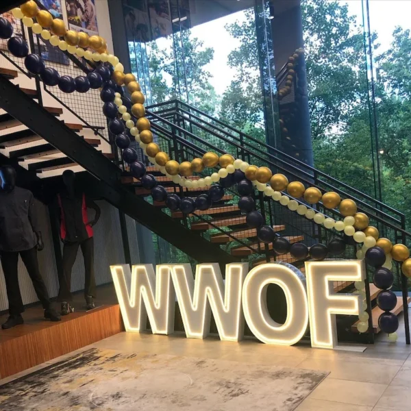 Large illuminated letters spelling "WWOF" placed near a staircase decorated with garlands of navy blue, gold, and white balloons.