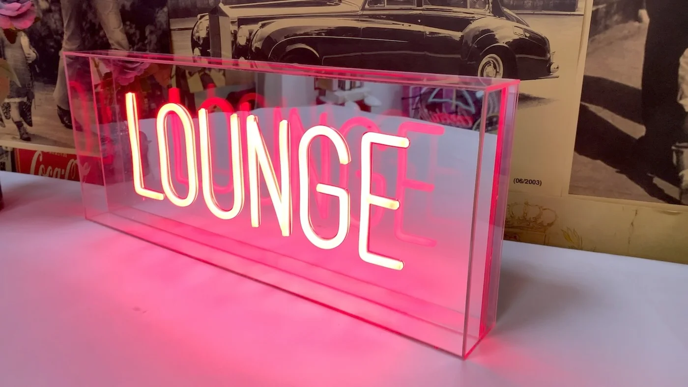 Neon sign with the word "LOUNGE" in pink letters, encased in a clear acrylic box, positioned on a table.