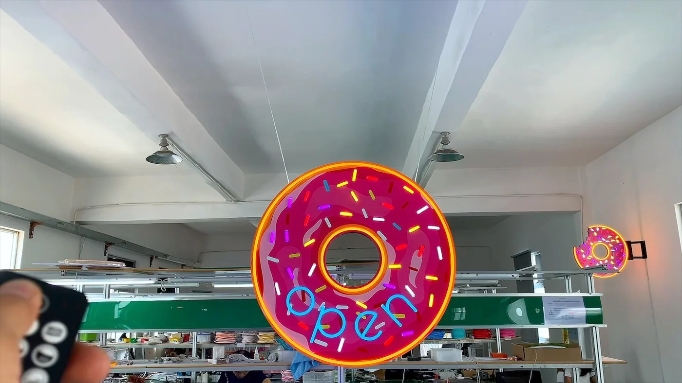 Neon sign in the shape of a pink frosted donut with colorful sprinkles, displaying the word "open" in blue letters. The sign is illuminated and hanging in a workshop environment, showcasing a vibrant and playful design ideal for a bakery or donut shop.