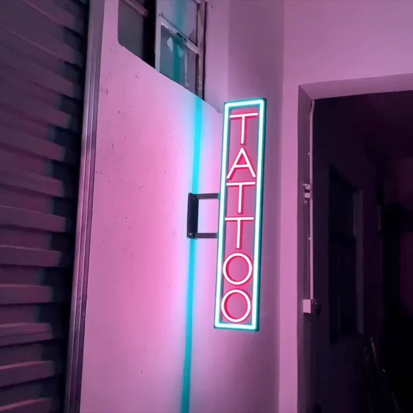 Vertical neon sign reading "TATTOO" in red letters, outlined with a blue glow, mounted on an exterior wall. The sign is illuminated against a pink and purple backdrop, creating a striking and edgy look suitable for a tattoo parlor.