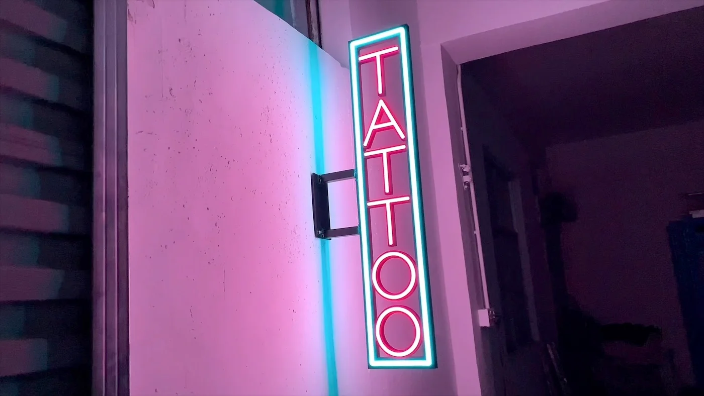 Vertical neon sign reading "TATTOO" in red letters, outlined with a blue glow, mounted on an exterior wall. The sign is illuminated against a pink and purple backdrop, creating a striking and edgy look suitable for a tattoo parlor.