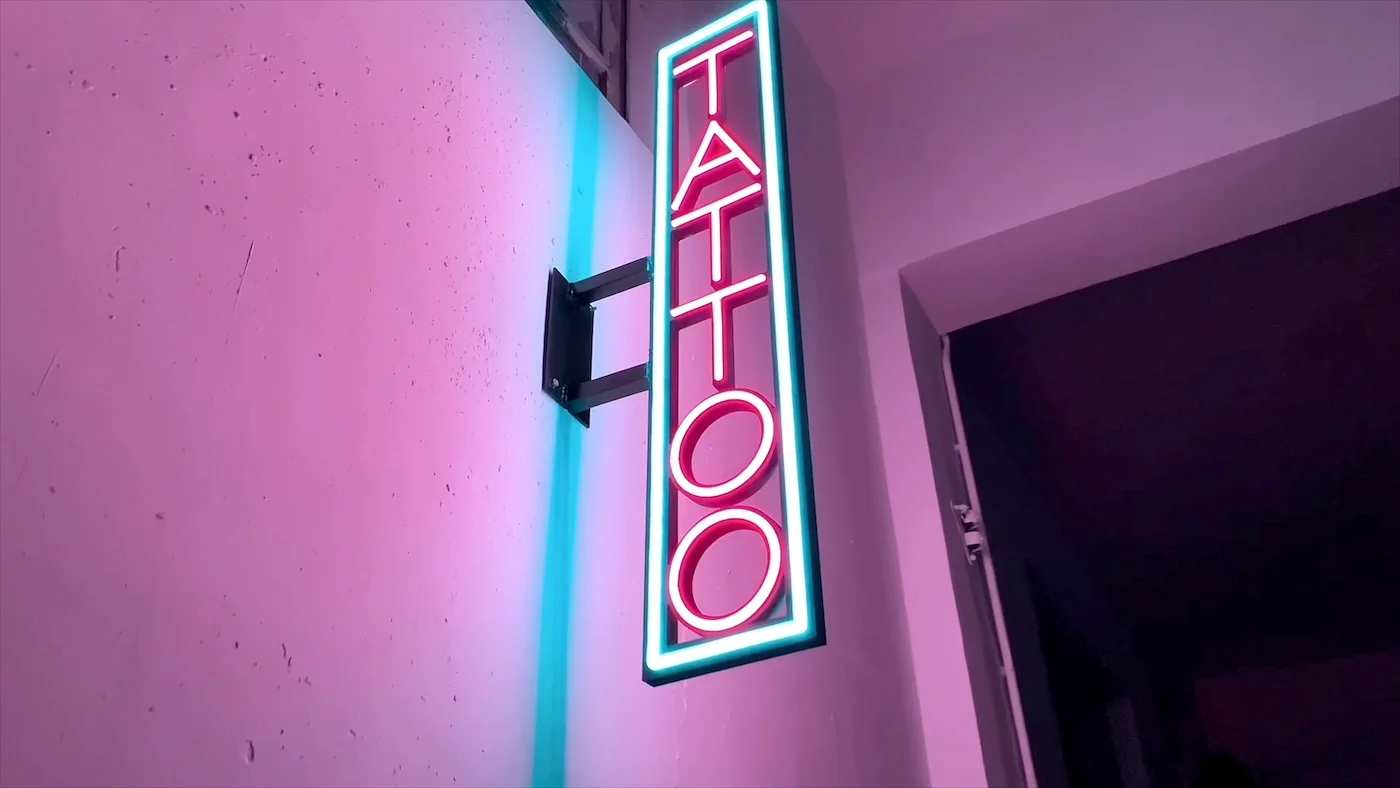 Vertical neon sign reading "TATTOO" in red letters, outlined with a blue glow, mounted on an exterior wall. The sign is illuminated against a pink and purple backdrop, creating a striking and edgy look suitable for a tattoo parlor.