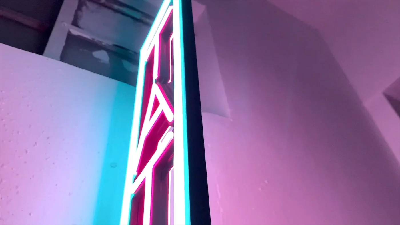 Vertical neon sign reading "TATTOO" in red letters, outlined with a blue glow, mounted on an exterior wall. The sign is illuminated against a pink and purple backdrop, creating a striking and edgy look suitable for a tattoo parlor.