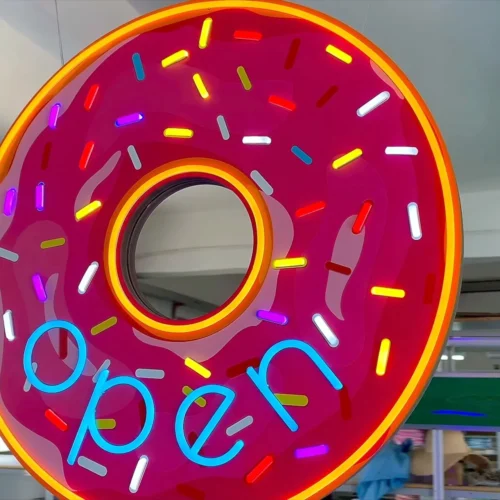 Neon sign in the shape of a pink frosted donut with colorful sprinkles, displaying the word "open" in blue letters. The sign is illuminated and hanging in a workshop environment, showcasing a vibrant and playful design ideal for a bakery or donut shop.