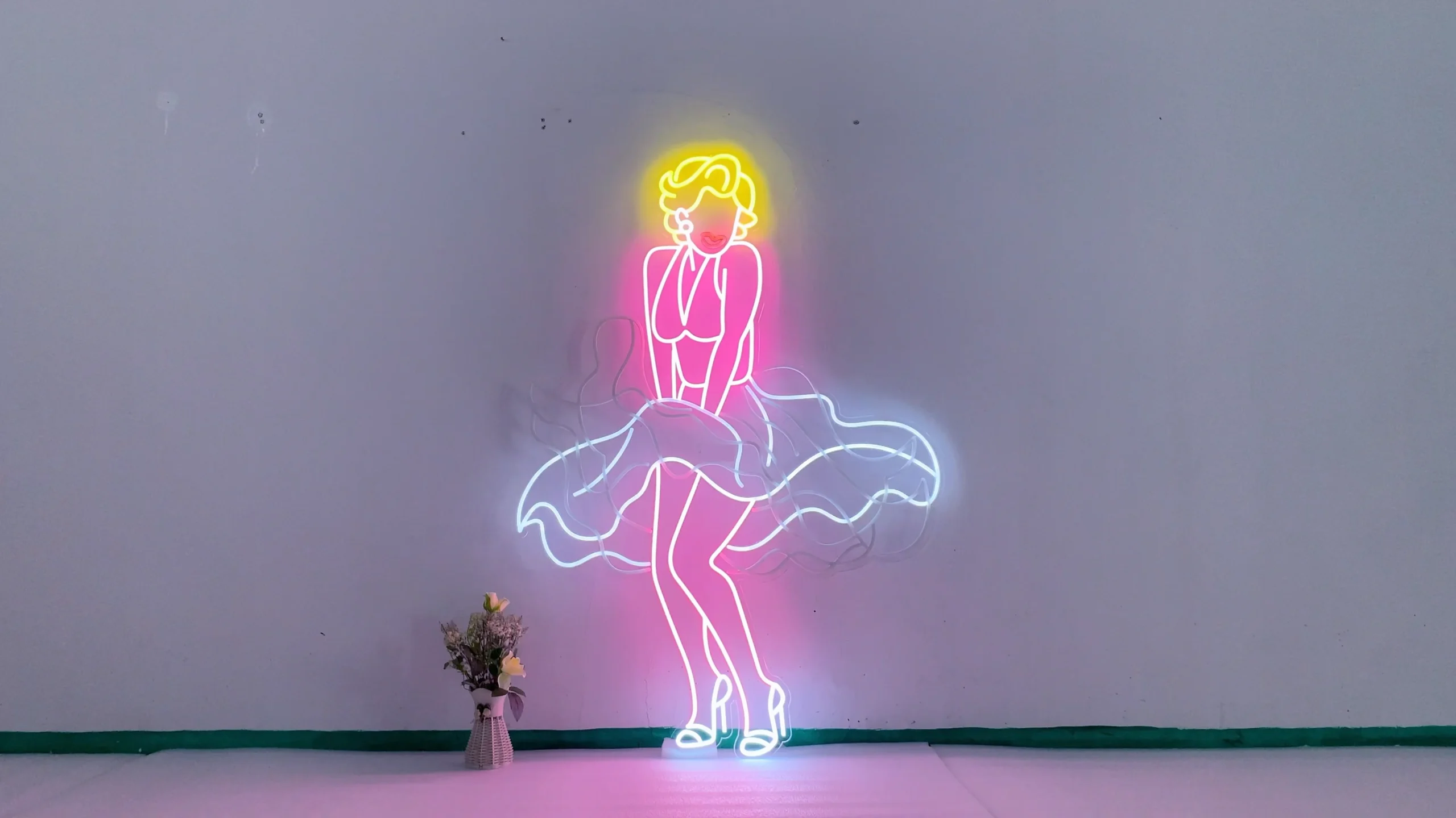 Neon sign depicting a stylized figure of Marilyn Monroe in her iconic pose with a flowing dress. The sign features bright colors, with yellow for her hair, pink for her dress and body, and white for the dress details and shoes.