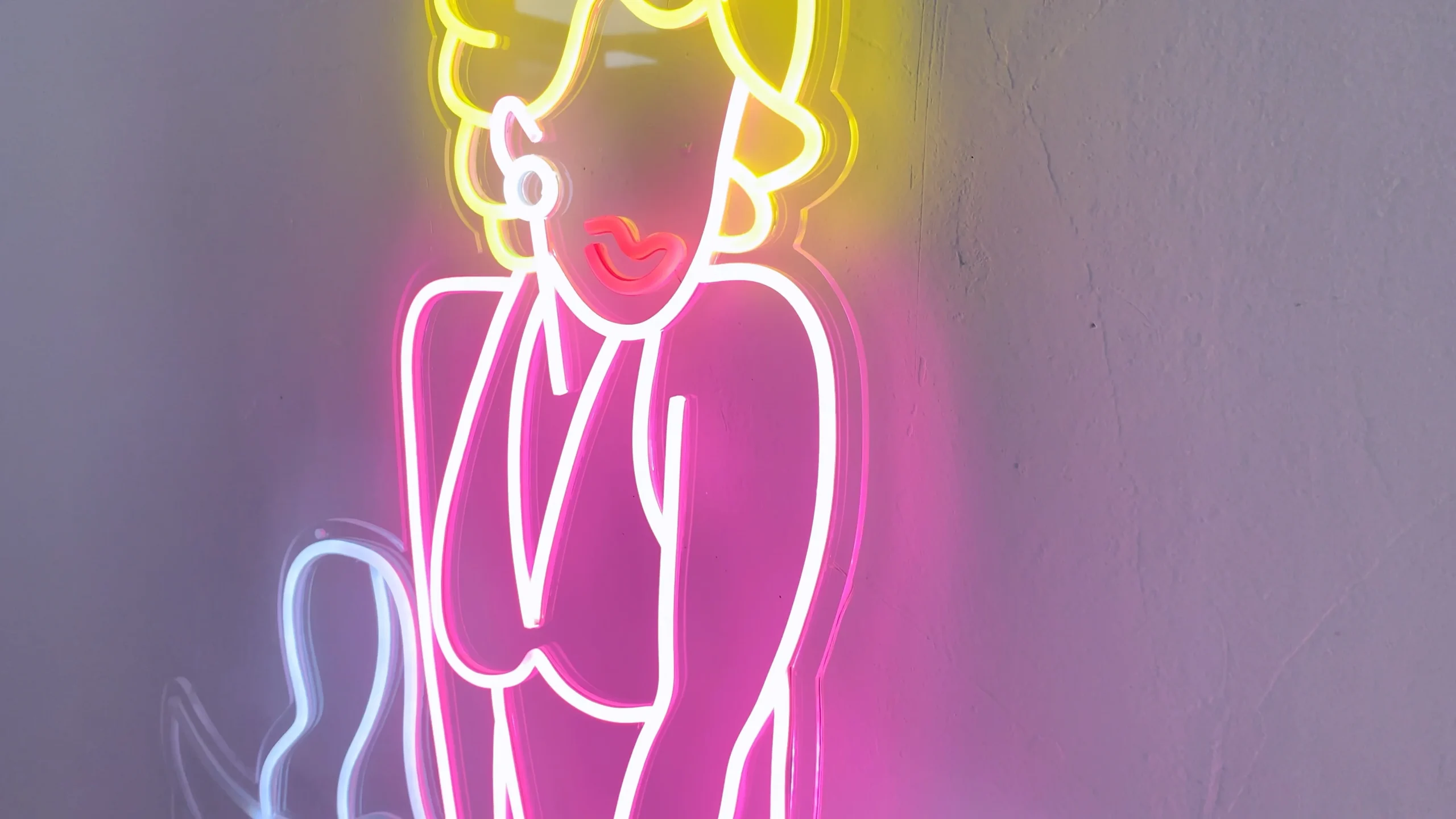 Neon sign depicting a stylized figure of Marilyn Monroe in her iconic pose with a flowing dress. The sign features bright colors, with yellow for her hair, pink for her dress and body, and white for the dress details and shoes.