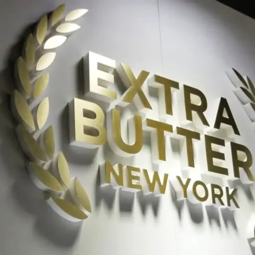 A 3D wall sign in gold letters reads "EXTRA BUTTER NEW YORK." The text is flanked by a laurel wreath design on both sides. The sign is mounted on a light-colored wall, with the letters casting shadows, creating a striking visual effect.