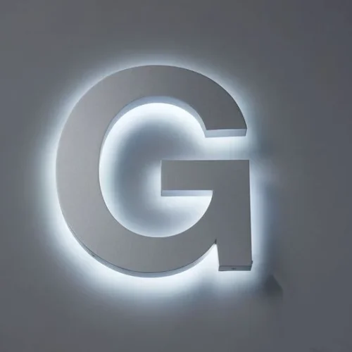 Illuminated letter sign mounted on a wall. The large, three-dimensional letter is backlit with a soft, white glow, creating a halo effect around the grey letter. The simple, bold design stands out against the wall, making it highly visible and modern.