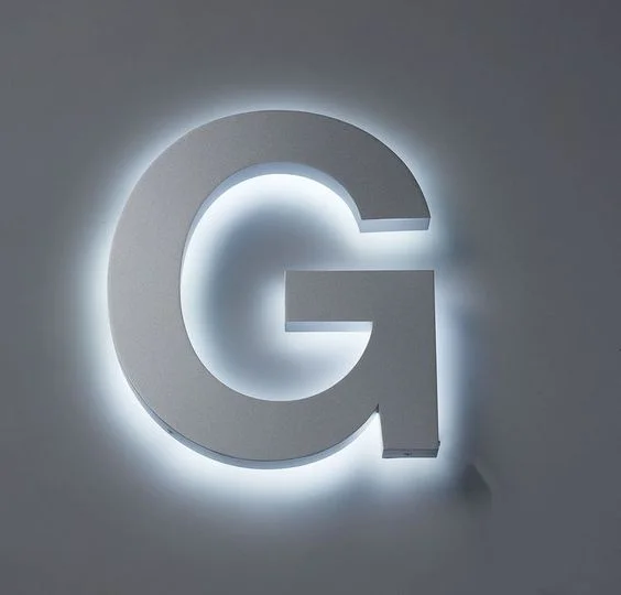 Illuminated letter sign mounted on a wall. The large, three-dimensional letter is backlit with a soft, white glow, creating a halo effect around the grey letter. The simple, bold design stands out against the wall, making it highly visible and modern.