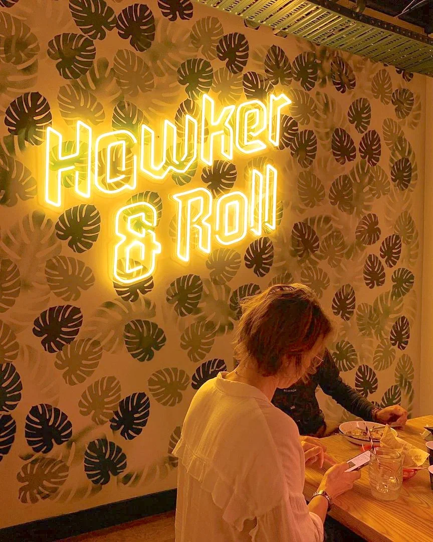 Neon sign with the words "Hawker & Roll" in yellow, mounted on a wall with a leafy pattern. The sign illuminates a cozy dining area.