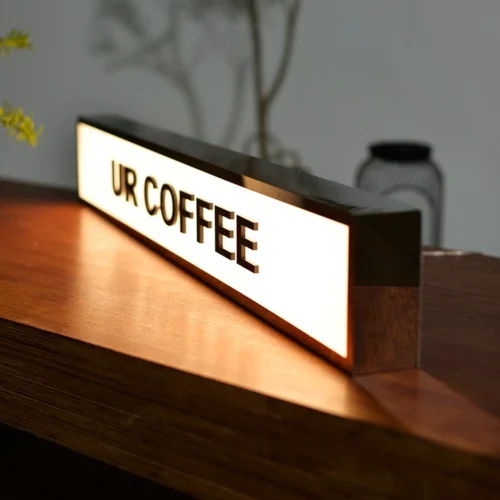 Illuminated "COFFEE" light box sign placed on a wooden surface, used as business signage for a coffee shop. The sign has a sleek black frame and emits a warm glow, creating a cozy ambiance.