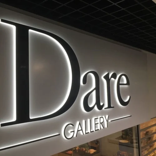 An illuminated wall sign for "Dare Gallery" features large black letters with a sleek, modern design. The word "Dare" is prominently displayed, with "GALLERY" in smaller letters below, both surrounded by horizontal lines. The sign is mounted on a light-colored wall with the letters backlit, creating a glowing effect.