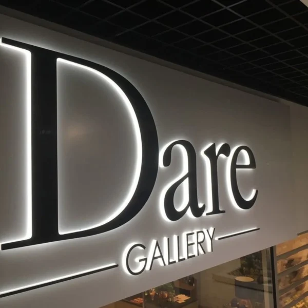 An illuminated wall sign for "Dare Gallery" features large black letters with a sleek, modern design. The word "Dare" is prominently displayed, with "GALLERY" in smaller letters below, both surrounded by horizontal lines. The sign is mounted on a light-colored wall with the letters backlit, creating a glowing effect.