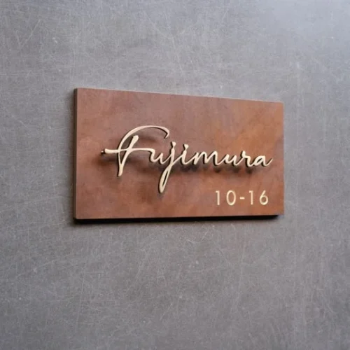 A rectangular brown metal sign with the name written in elegant, cursive letters. Below the name, the numbers "10-16" are displayed. The sign is mounted on a textured gray wall. This signage is suitable for business or home identification.