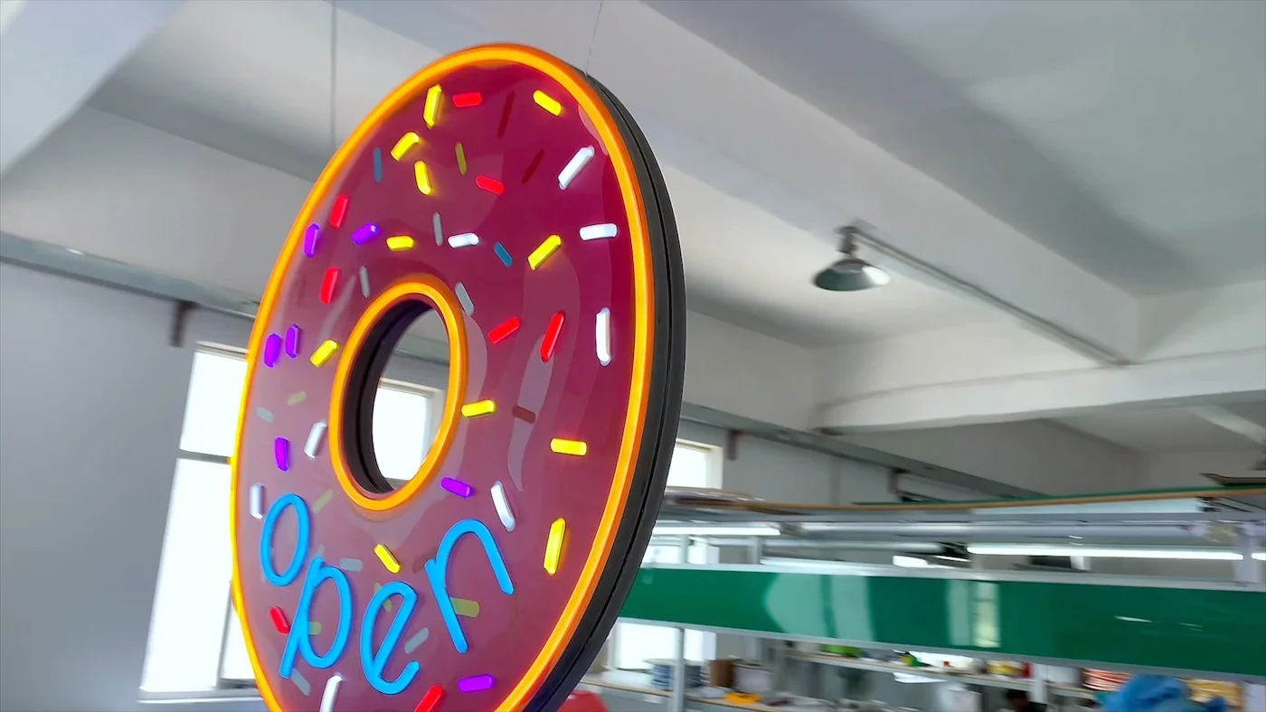 Neon sign in the shape of a pink frosted donut with colorful sprinkles, displaying the word "open" in blue letters. The sign is illuminated and hanging in a workshop environment, showcasing a vibrant and playful design ideal for a bakery or donut shop.