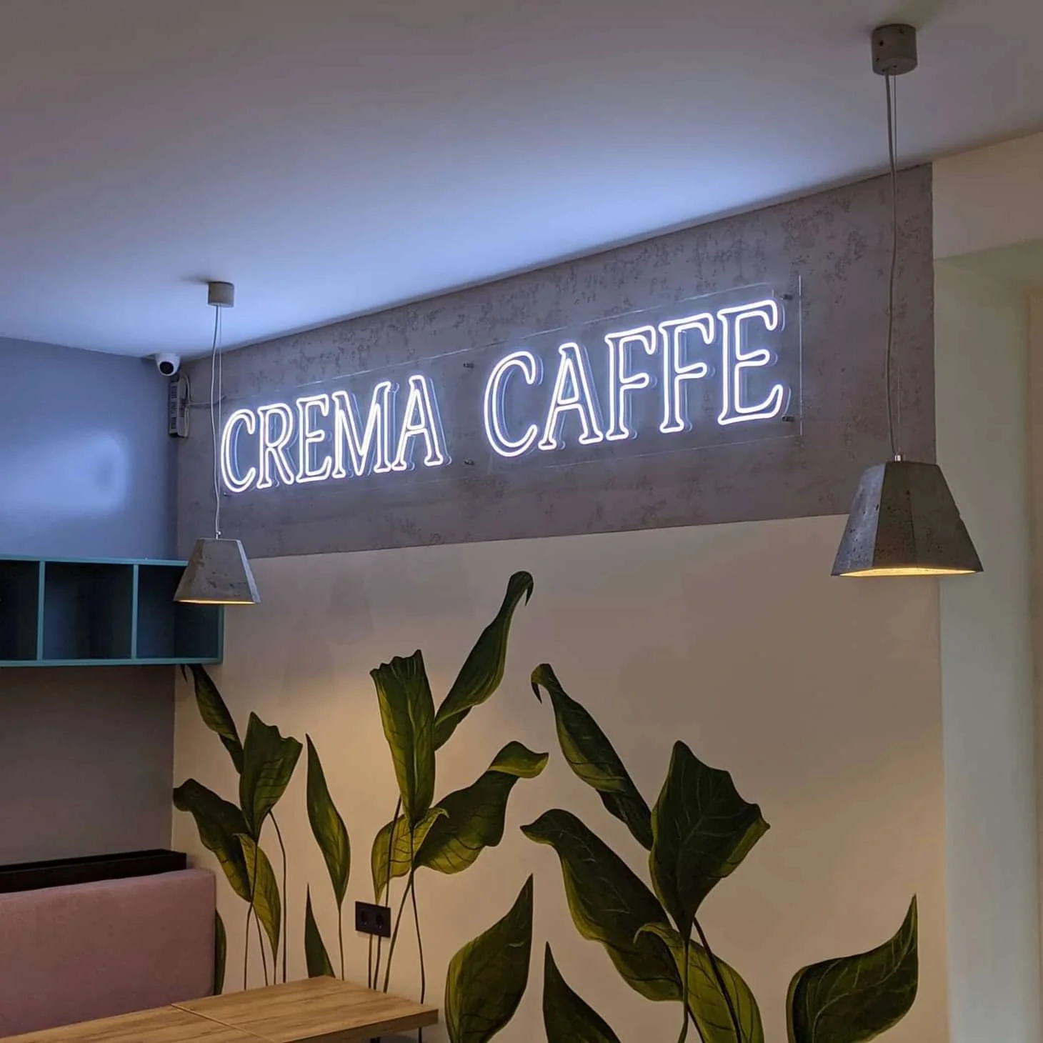 Neon sign reading "CREMA CAFFE" in white, mounted on a light gray wall above a seating area in a café.