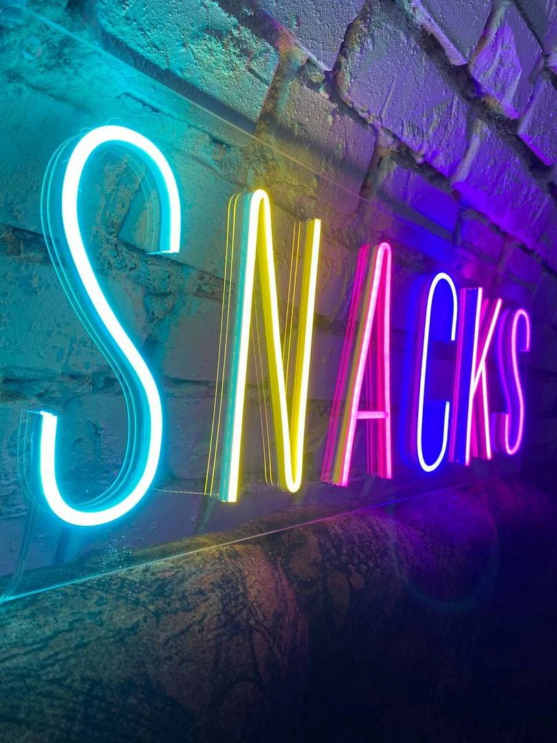 Vibrant neon sign displaying the word "SNACKS" in various colors, mounted on a textured brick wall. The letters are illuminated in blue, yellow, pink, and green, creating a fun and eye-catching effect suitable for a snack bar or casual dining spot.