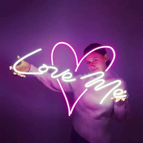 Woman holding a neon sign with the words "Love Me" in white script and a pink heart outline, against a purple background. The sign creates a romantic and whimsical vibe, perfect for events or personal decoration.