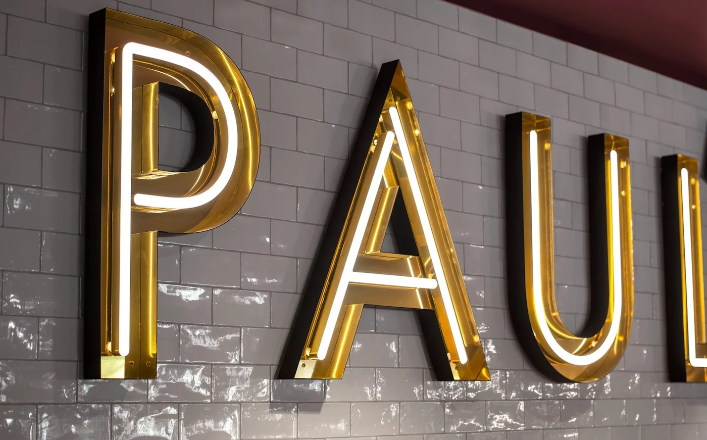 Illuminated neon sign reading "PAUL'S" mounted on a tiled wall inside a restaurant. The sign features large, bold letters with a yellow and white glow, enhancing the upscale ambiance of the venue. The sign is situated above a hallway leading to other areas of the restaurant.