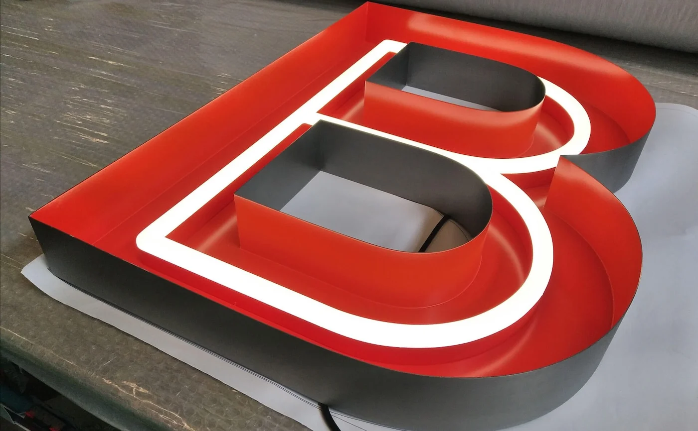 Close-up of a large letter "B" in red with a white neon outline, lying flat on a workbench. The letter features a bold and modern design, highlighting the craftsmanship involved in creating custom neon signage.