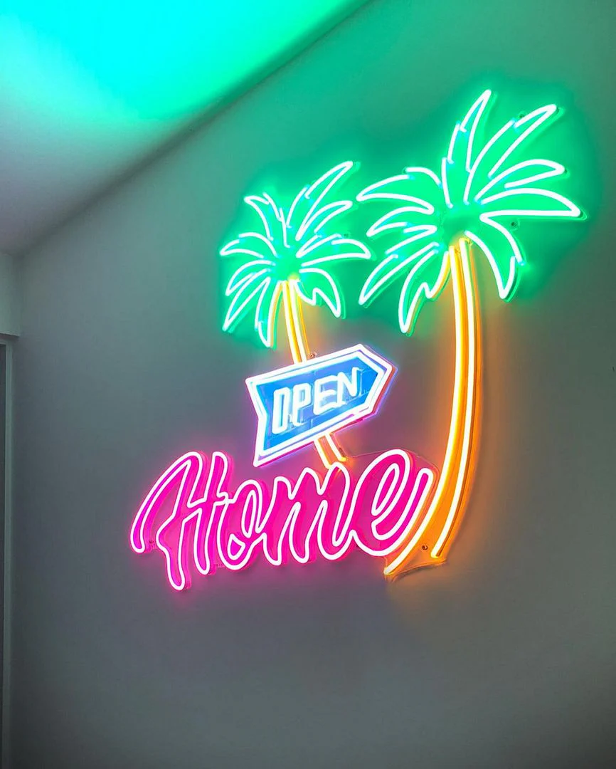 Colorful neon sign with two palm trees, a blue "OPEN" sign, and the word "Home" in pink script, mounted on a wall. The sign emits a tropical and welcoming glow, ideal for a beach-themed venue or home decor.