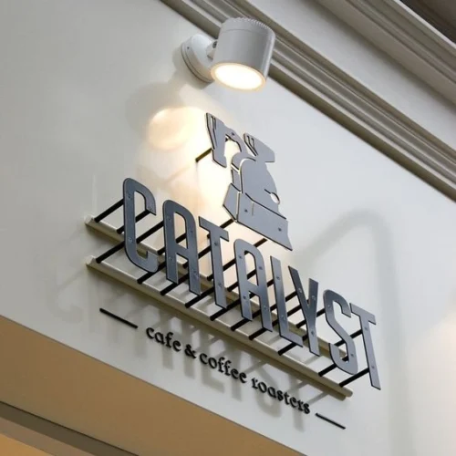 A modern sign for "Catalyst" cafe and coffee roasters, mounted on a light-colored wall. The sign features bold, metallic letters spelling "Catalyst" above the phrase "cafe & coffee roasters" in smaller text. Above "Catalyst," there is an icon of a coffee roaster. The sign is illuminated by a white spotlight, adding a sleek and professional appearance.