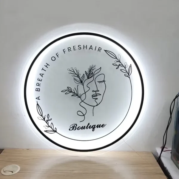 A round illuminated sign features the text "A BREATH OF FRESHAIR" along the top and "Boutique" in cursive at the bottom. In the center, there is a minimalist line drawing of a face adorned with leaves and floral elements. The sign is mounted on a plain white wall above a light-colored wooden surface.