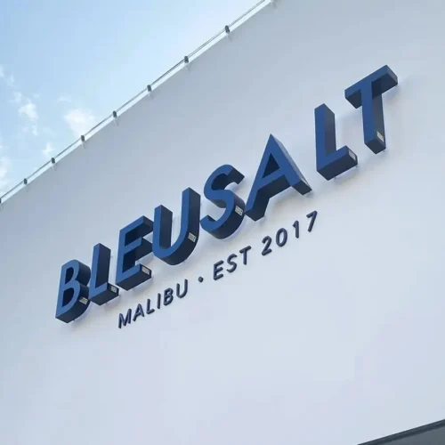 Blue 3D signage on a white building facade. The sign is prominently displayed under a clear blue sky.