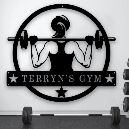 A black metal wall sign features a silhouette of a muscular person lifting a barbell. The figure is enclosed in a circular frame with the text "NAME'S GYM" below, flanked by stars. The sign is mounted on a light-colored wall in a gym setting, with a barbell and weight rack visible to the right.