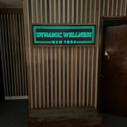 A neon sign mounted on a striped wooden wall reads "DYNAMIC WELLNESS" in large green letters with "NEW YORK" below, also in green. The sign is framed in wood and positioned above a wooden bench. A dark-colored door is visible to the right.