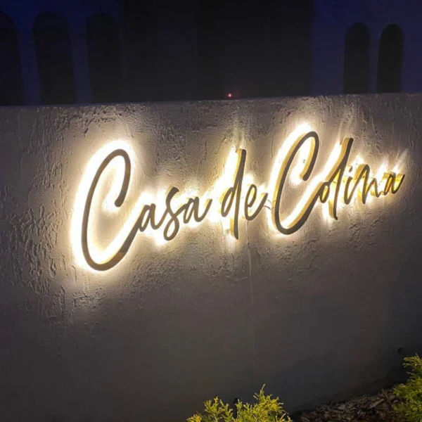 Illuminated "Casa de Colina" sign mounted on a textured wall. The sign features cursive lettering in a warm, glowing light, creating a welcoming ambiance. The lighting accentuates the elegant script, making it stand out against the dark background, perfect for evening visibility.