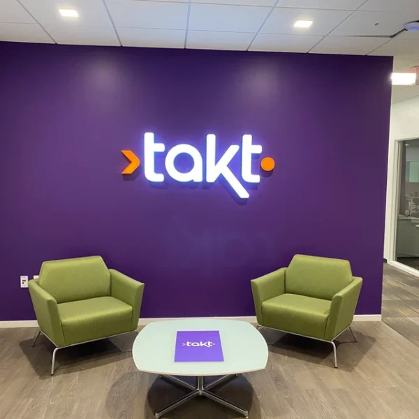 A modern office lounge area features a purple accent wall with an illuminated sign displaying the logo "takt." in white letters with an orange arrow and dot.