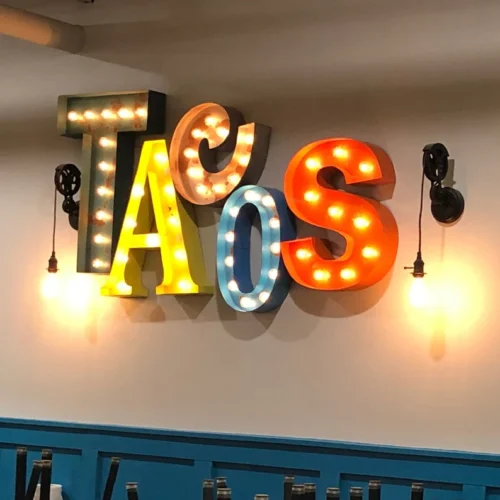 A colorful wall sign spells out "TACOS" with each letter in a different vibrant color and style, outlined with lightbulbs. The letters are mounted on a light-colored wall.