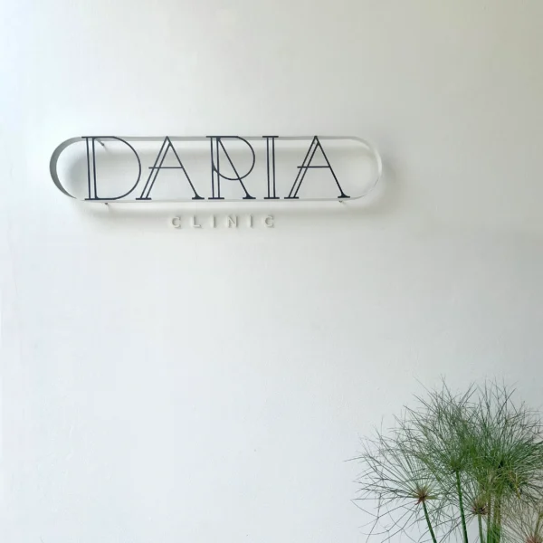 Minimalist sign mounted on a white wall. The sign features elegant, thin black letters inside an elongated oval shape, with name in a large, stylish font and "Clinic" in smaller, spaced-out letters below. The clean and modern design complements the simple wall, with a green plant adding a touch of nature to the setting.