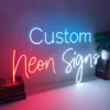 Neon sign displaying the words "Custom Neon Signs" mounted on a plain wall. The word "Custom" is in blue, "Neon" in red, and "Signs" in white.