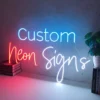 Neon sign displaying the words "Custom Neon Signs" mounted on a plain wall. The word "Custom" is in blue, "Neon" in red, and "Signs" in white.