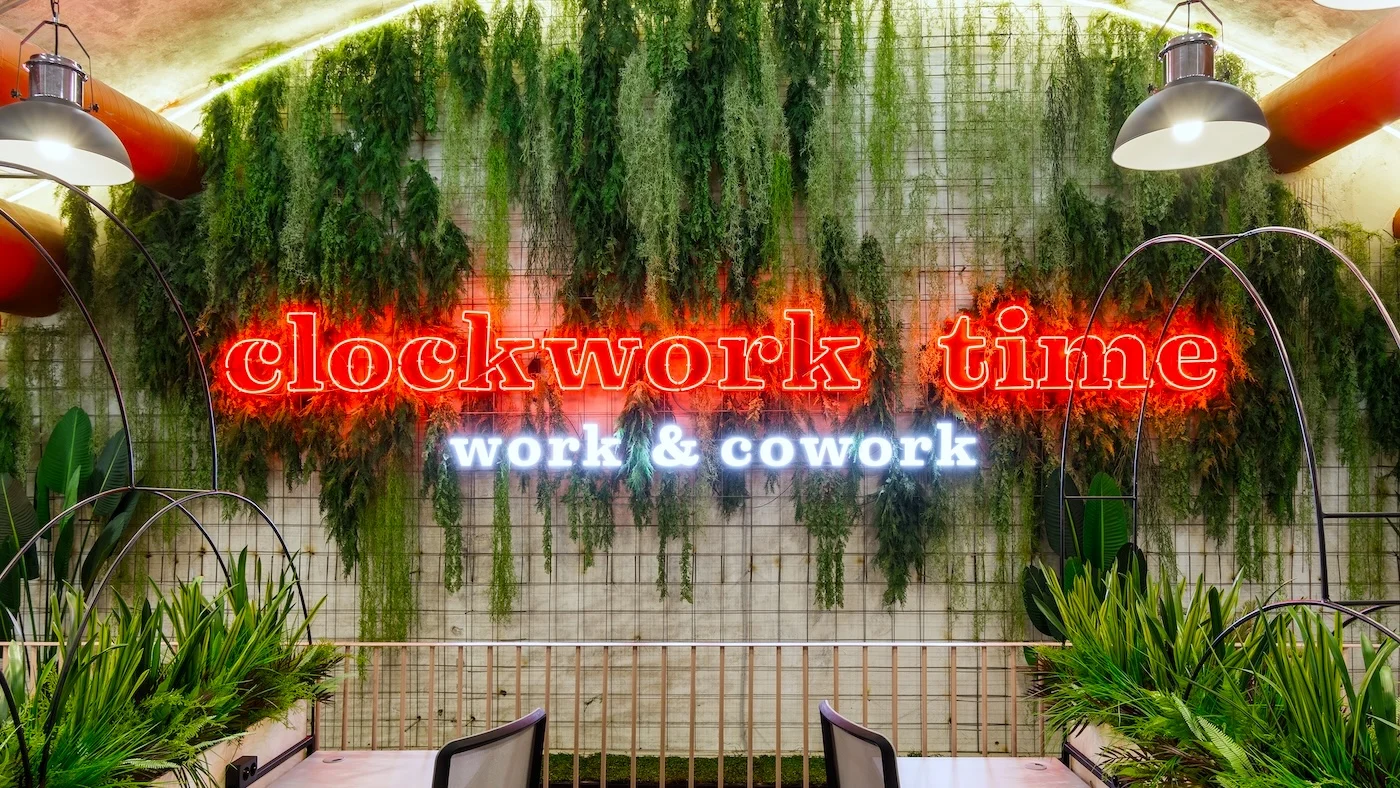 Vibrant neon sign reading "clockwork time work & cowork" mounted on a wall covered with lush greenery in a coworking space. The words "clockwork time" are in red, while "work & cowork" are in white, creating a striking visual contrast.