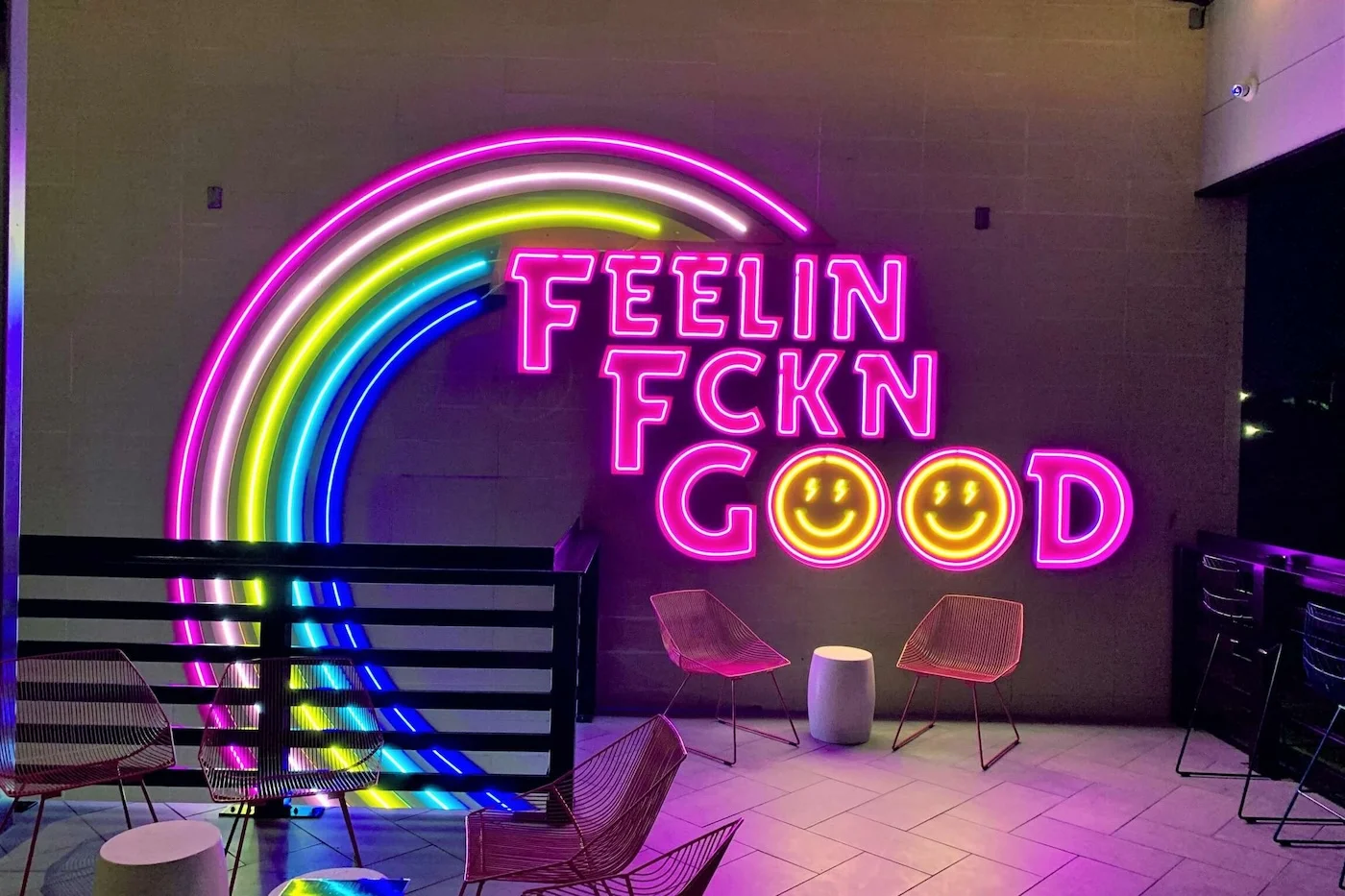 Bright neon sign featuring the phrase "FEELIN FCKN GOOD" with two smiley faces, mounted on an indoor wall. The words are in pink, and the sign is complemented by a colorful neon rainbow arc.