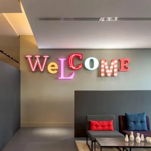 A decorative wall sign in a modern lounge spells out "WELCOME" with each letter in a different style and color. The letters are a mix of red, pink, yellow, green, and blue, with some featuring lightbulbs. The sign is mounted on a neutral-colored wall.