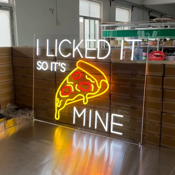 A colorful neon sign on a table features the text "I LICKED IT SO IT'S MINE" in white letters. Below the text is a glowing yellow and red outline of a slice of pizza with pepperoni.