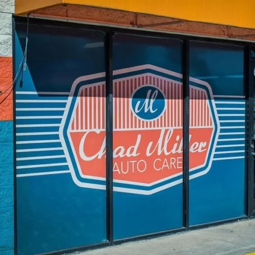 A storefront window sign for "Chad Miller Auto Care" features a retro design with a red and blue color scheme. The logo consists of a hexagonal shape with a stylized "M" at the top and the business name in cursive script below. The background has horizontal white stripes and is framed by blue and red sections of the building.