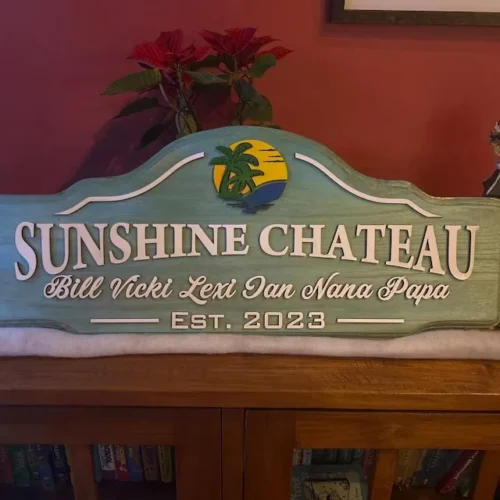 A wooden sign with a curved top reads "SUNSHINE CHATEAU" in large white letters. Below, it lists names: "Bill Vicki Lexi Ian Nana Papa" in a cursive font. The sign also displays "Est. 2023" at the bottom. The top features a colorful emblem of a palm tree against a setting sun. The sign is placed indoors against a red wall with potted plants visible in the background.