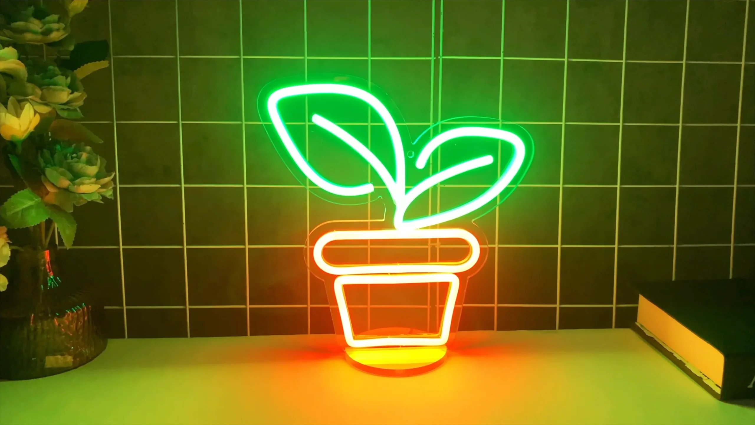 A desktop neon sign depicts a green-leaf plant and an orange flowerpot. The sign is placed on a desk, emitting vibrant light, and can be dimmed. It can also serve as a night light and an ambient light.